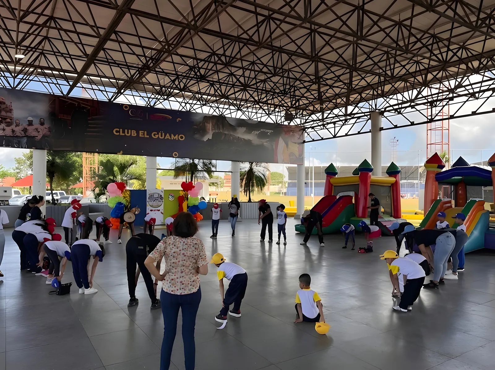 Niños con franela blanca y gorra amarilla hacen ejercicios guiados en una cancha techada, con castillos inflables al fondo.