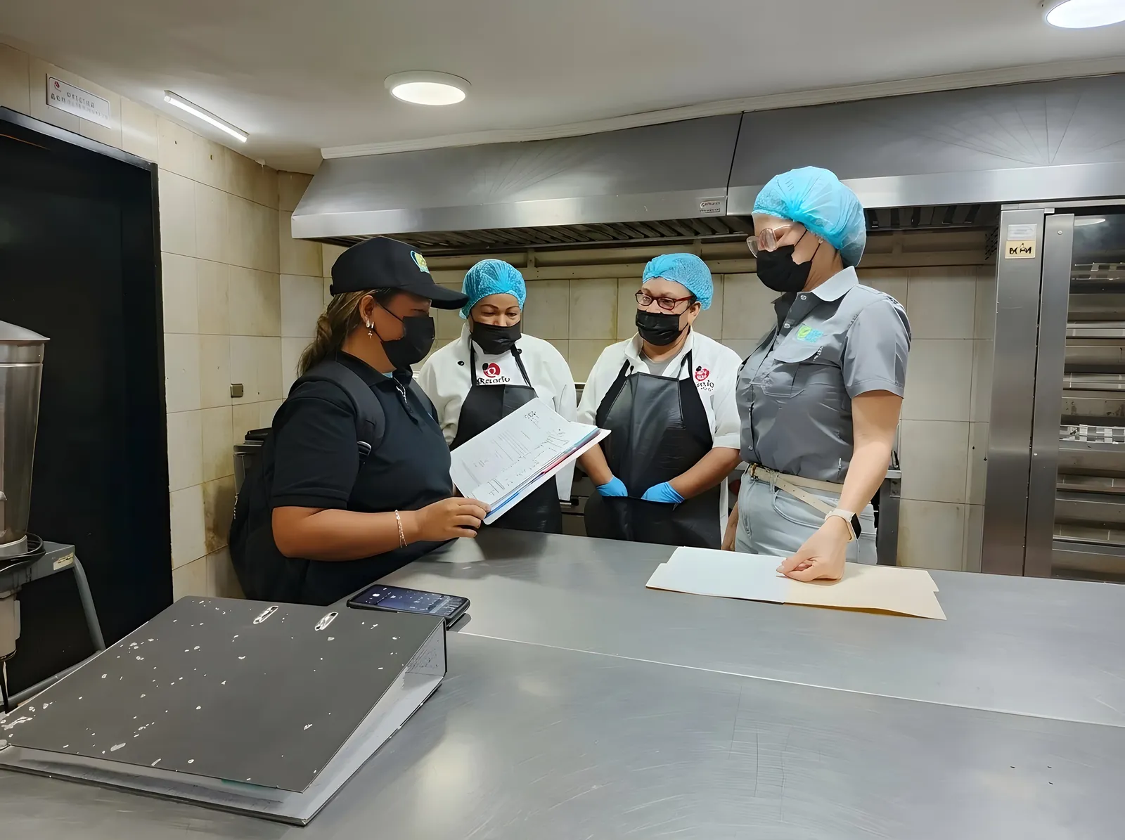 Supervisoras de BELT y cocineras con gorro y tapabocas revisan una carpeta en la cocina.