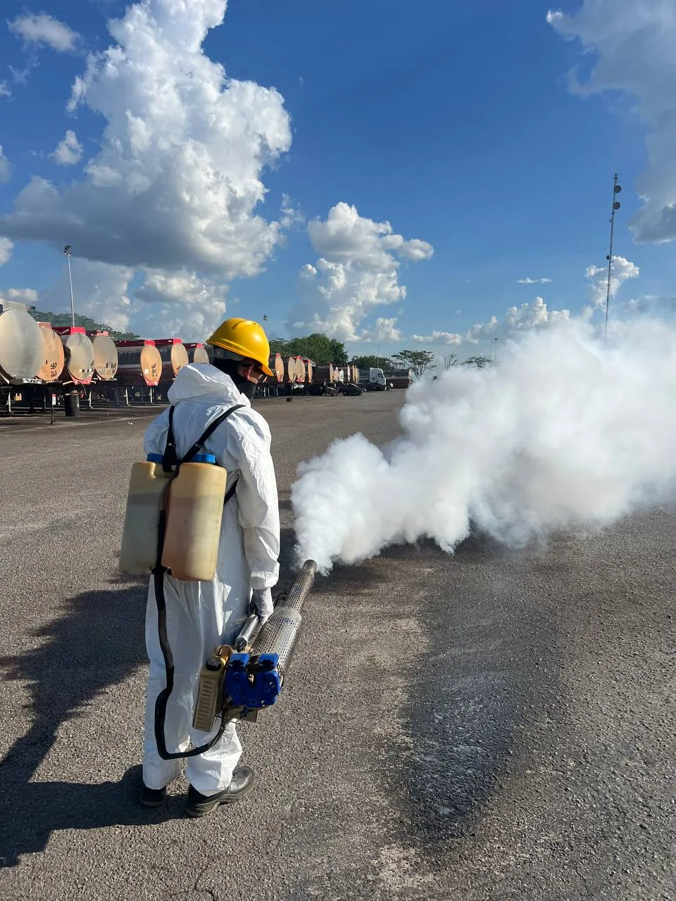 Operario de espaldas con la termonebulizadora y una nube blanca sobre el asfalto; al fondo, la fila de cisternas.