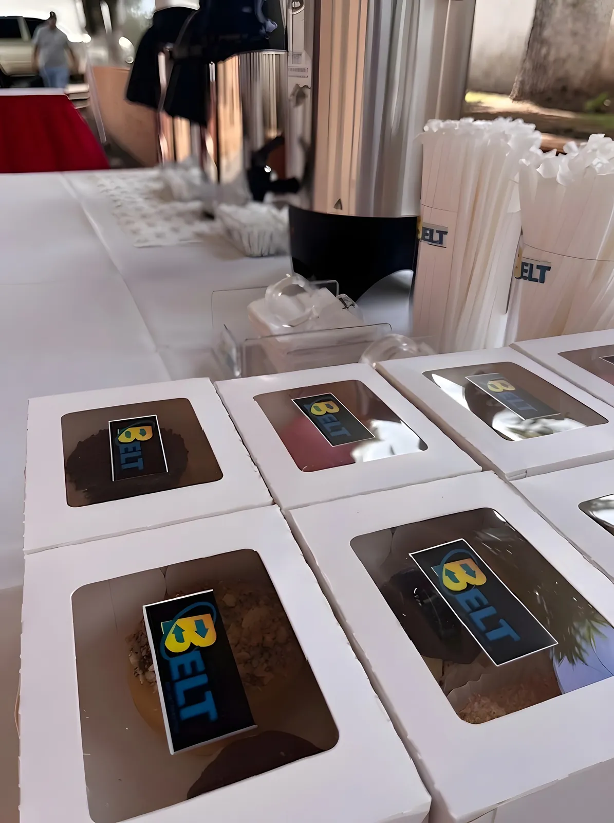 Postres en cajas blancas con ventana y etiqueta de BELT, frente a los termos de café.